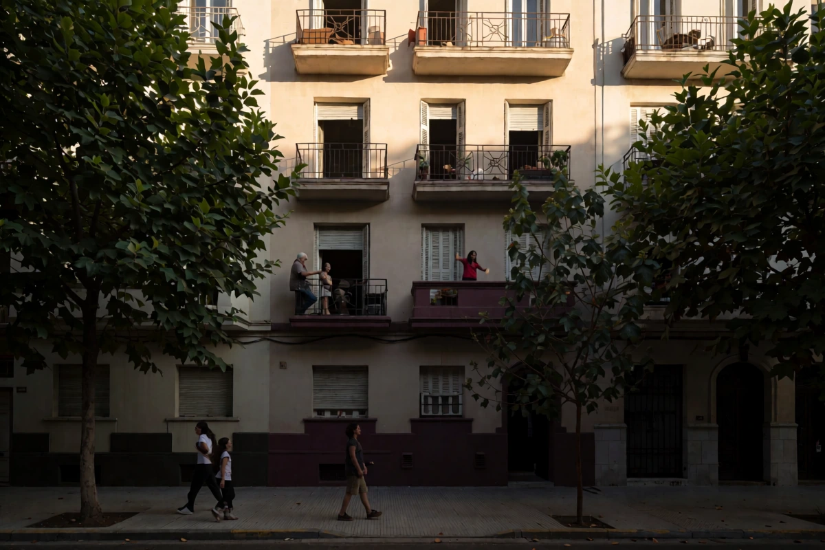 A sunlit residential street with apartment balconies, tree shadows, and three pedestrians